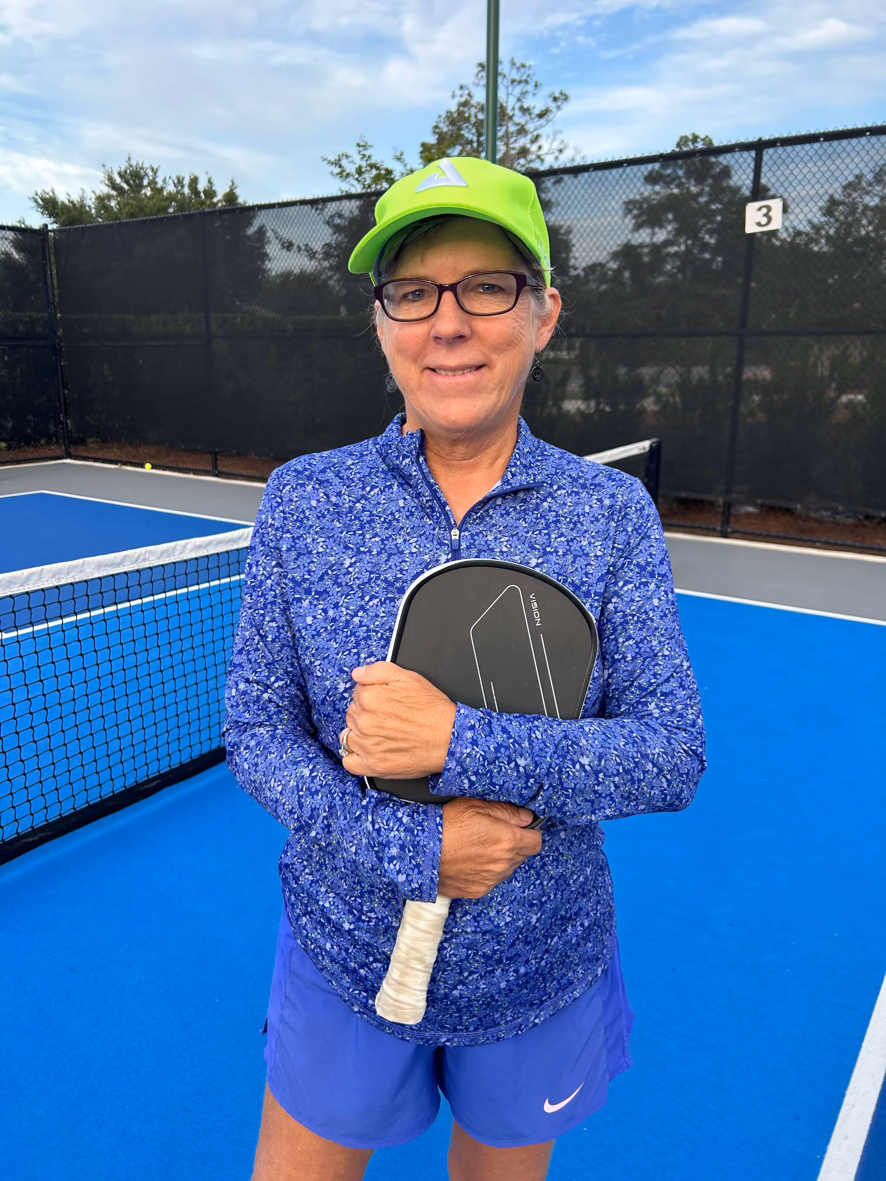 Woman in a neon green cap, glasses, and blue floral jacket holding a black pickleball paddle on a blue pickleball court.