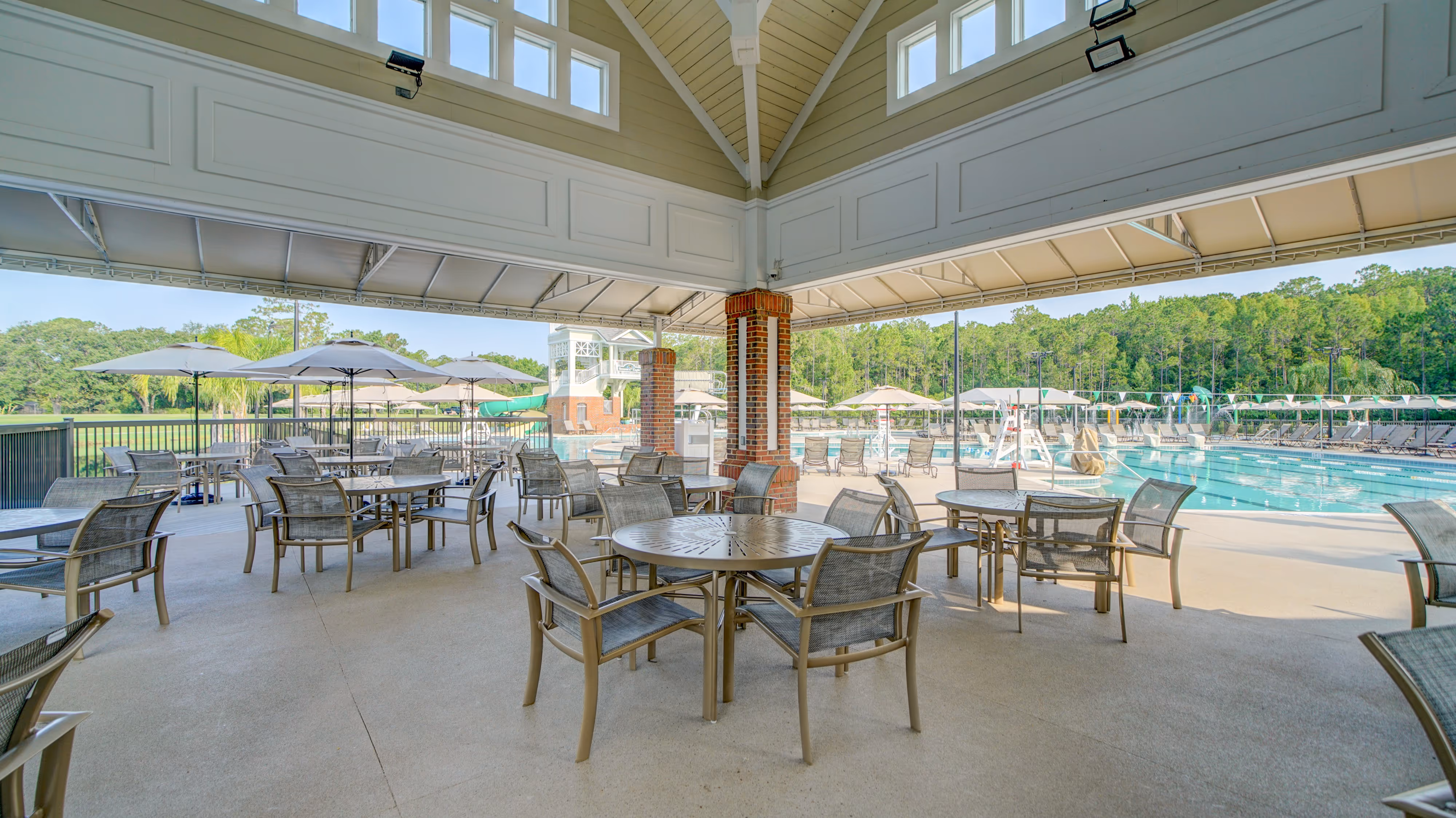 Covered patio area with round metal tables and mesh chairs overlooking a swimming pool and lounge chairs with umbrellas.