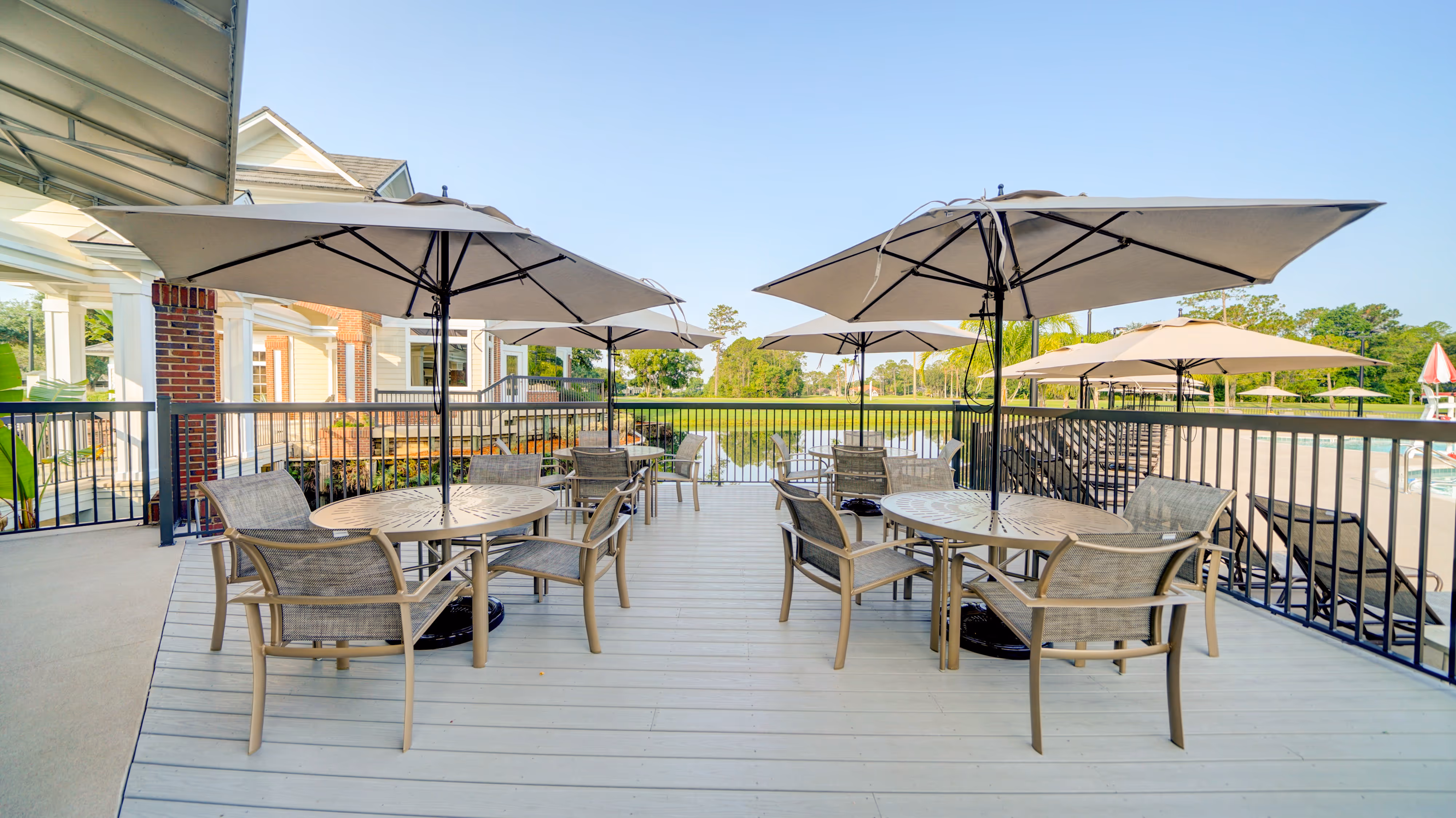Outdoor patio with round tables, chairs, and large beige umbrellas overlooking a pond and green landscape.