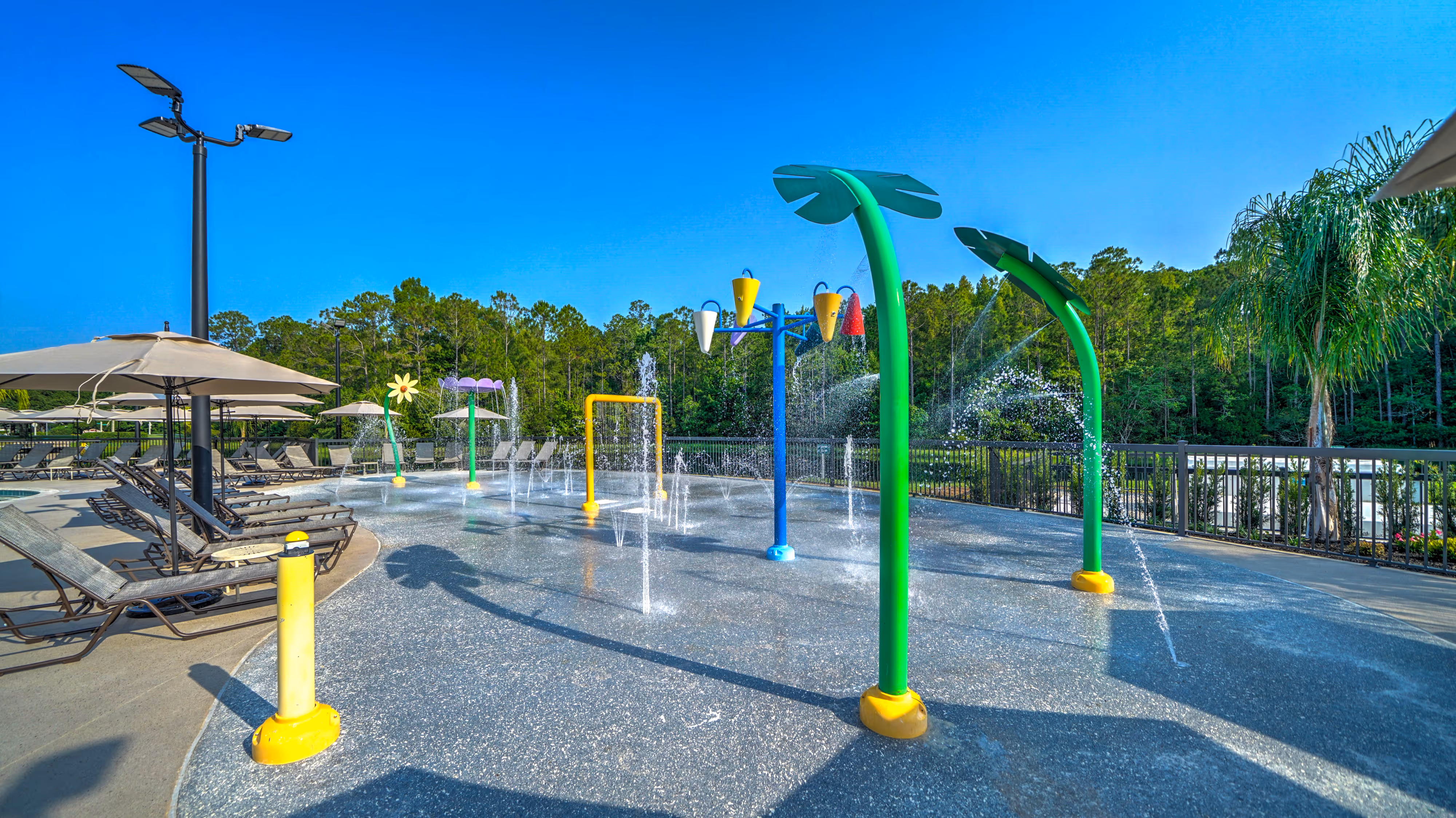 Splash pad with colorful water play structures shaped like leaves, flowers, and cones, surrounded by lounge chairs and umbrellas under a clear blue sky.