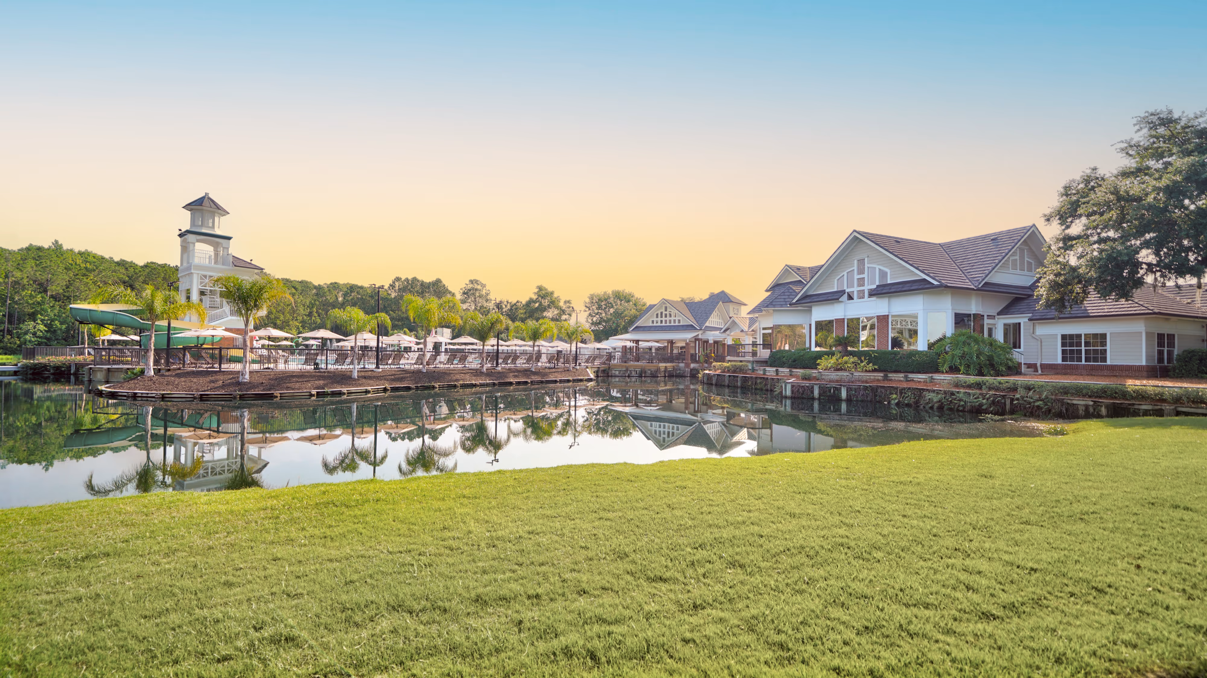 Lakefront resort with grassy shore, wooden dock, waterslide tower, and several houses against a clear sky at sunset.