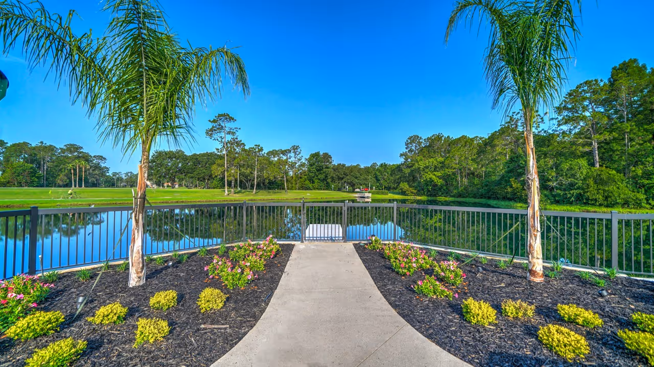 Concrete path bordered by flower beds and palm trees leading to a gated dock on a calm lake with trees and blue sky in the background.