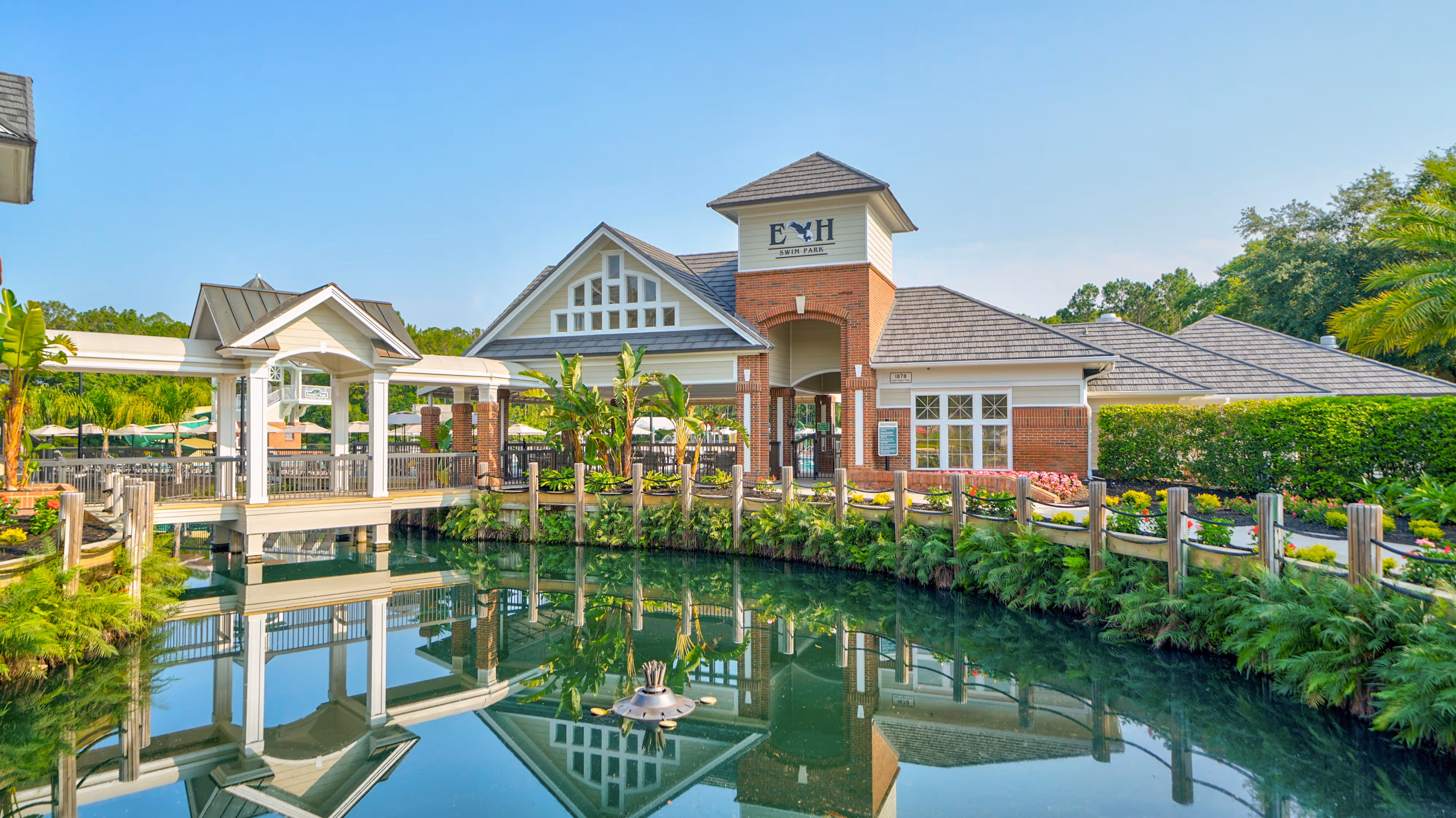 Swim park entrance building with a covered walkway over calm water reflecting the structure, surrounded by greenery and clear blue sky.