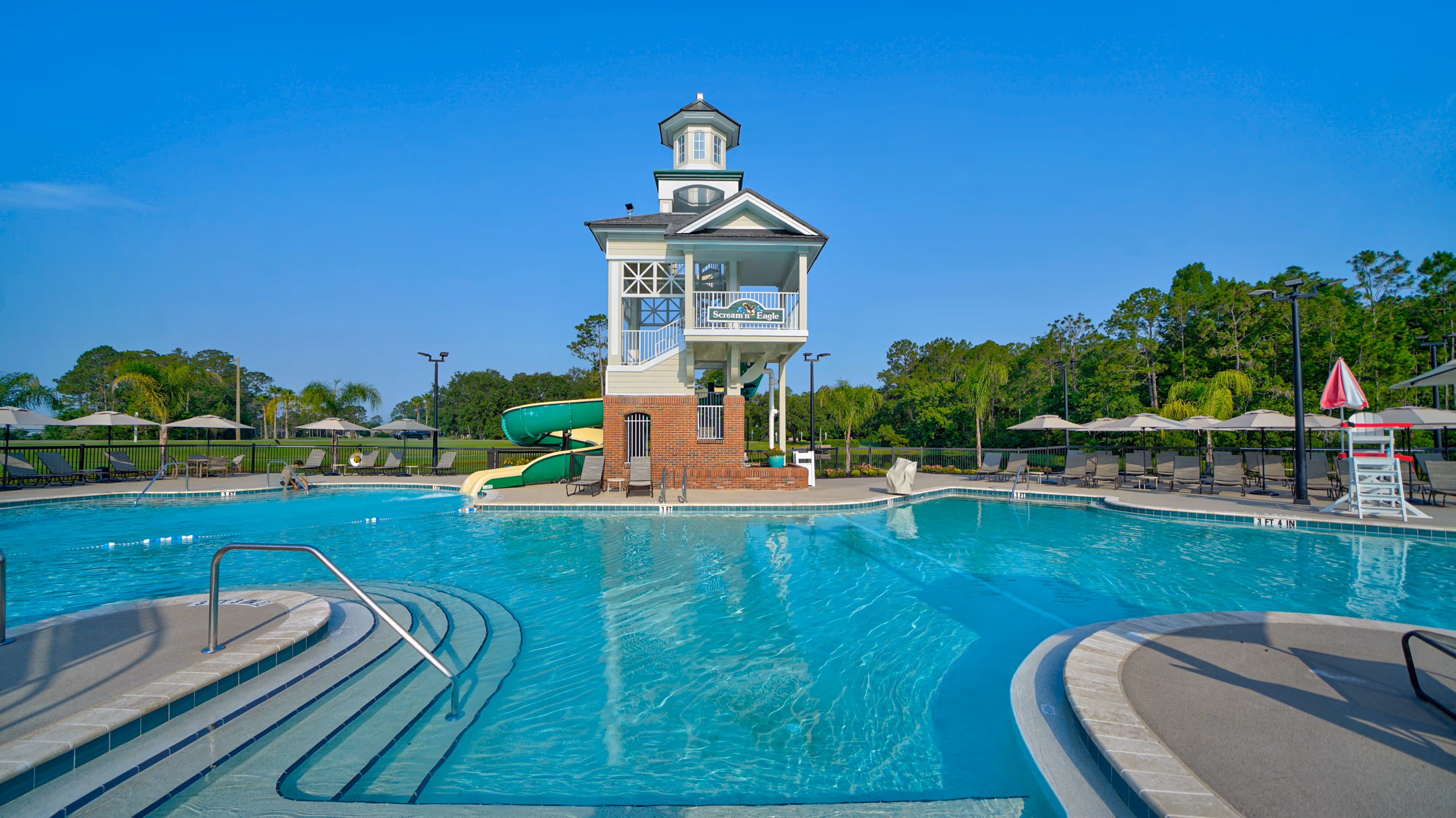 Outdoor swimming pool with a multi-story lifeguard tower and green water slide under clear blue sky.
