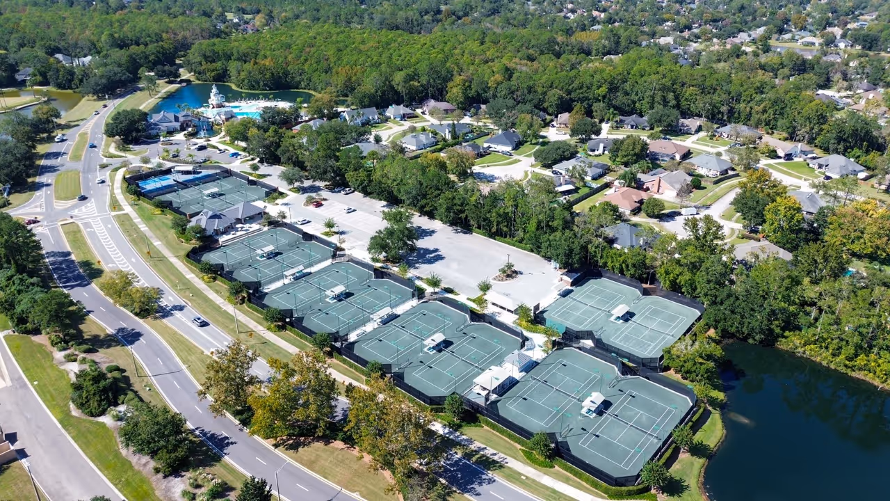 Aerial view of a suburban neighborhood featuring multiple tennis courts, a swimming pool area, roads, and surrounding greenery.