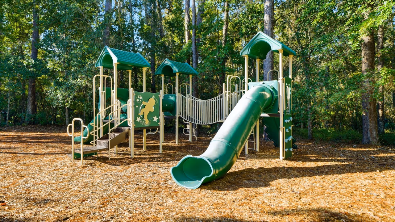 Green playground structure with slides, climbing wall, and rope bridge surrounded by trees and covered with wood chips.