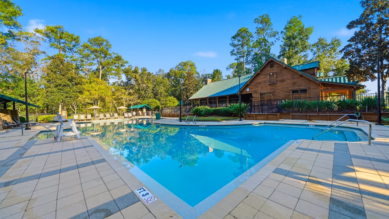 Outdoor swimming pool with clear blue water surrounded by lounge chairs and a wooden building with a green roof in the background under a bright blue sky.