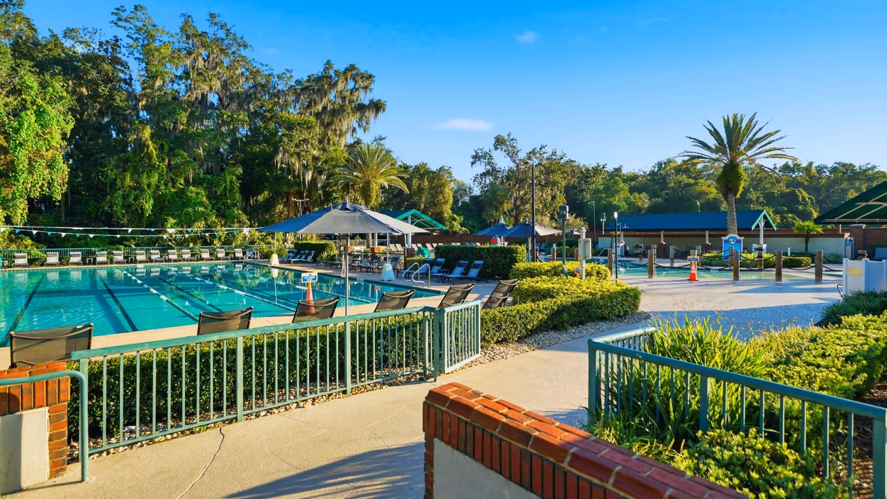 Outdoor community pool with lounge chairs, umbrellas, and surrounding greenery under a clear blue sky.