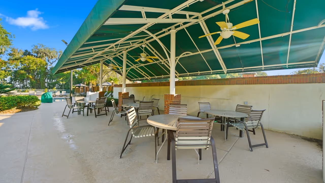 Outdoor patio area with multiple round tables and chairs under a green canopy with ceiling fans.