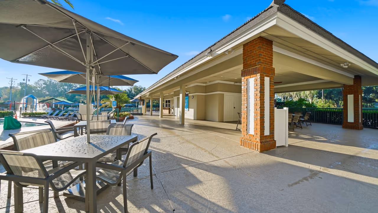 Outdoor poolside area with tables, chairs, umbrellas, and a covered pavilion with brick columns.