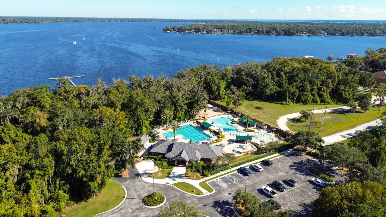 Aerial view of a lakeside community featuring a large swimming pool, clubhouse, parking lot, and lush green trees by a calm blue lake.