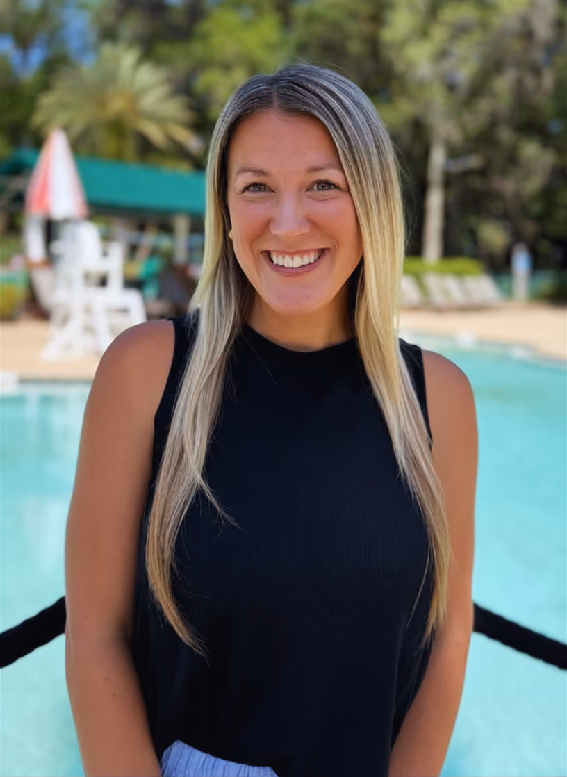 Smiling woman with long blonde hair wearing a black sleeveless top standing by a pool outdoors.