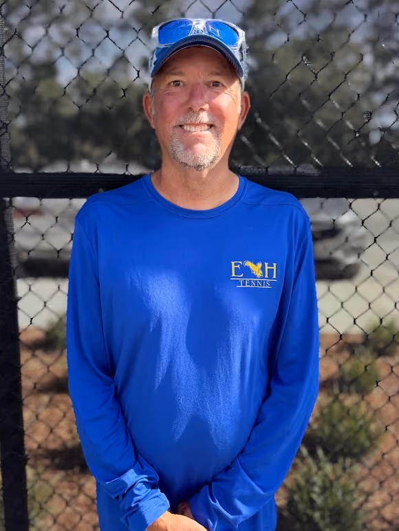 Man in blue long-sleeve shirt and cap with sunglasses stands in front of a tennis court fence.