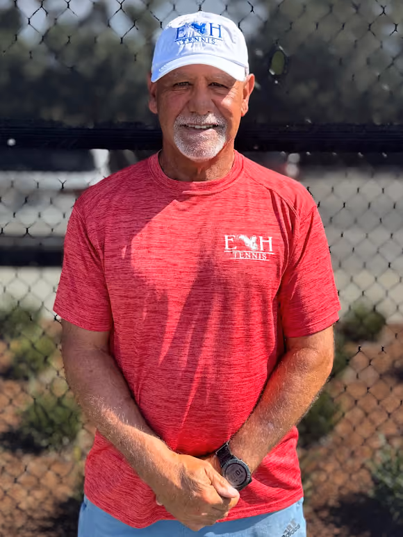 Older man in a white EH Tennis cap and red shirt standing in front of a chain-link fence.