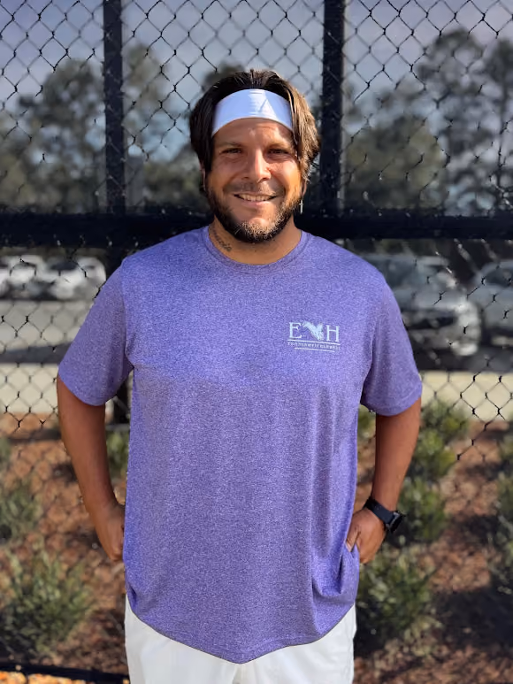 Smiling man with white headband and purple shirt standing in front of a chain-link fence outdoors.
