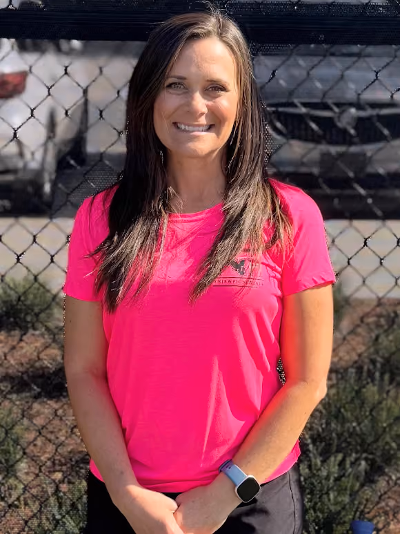 Smiling woman with long brown hair wearing a bright pink shirt and smartwatch, standing by a chain-link fence.