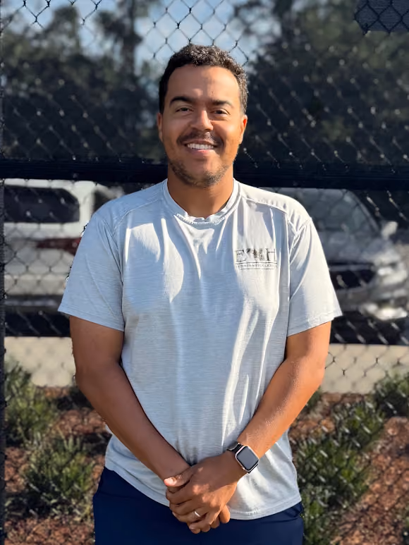 Smiling man with short curly hair wearing a light gray shirt and smartwatch standing by a chain-link fence.