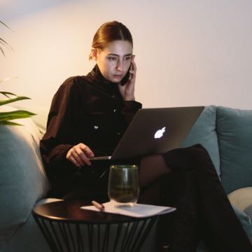 Someone sitting on a couch with a laptop and on a phone.
