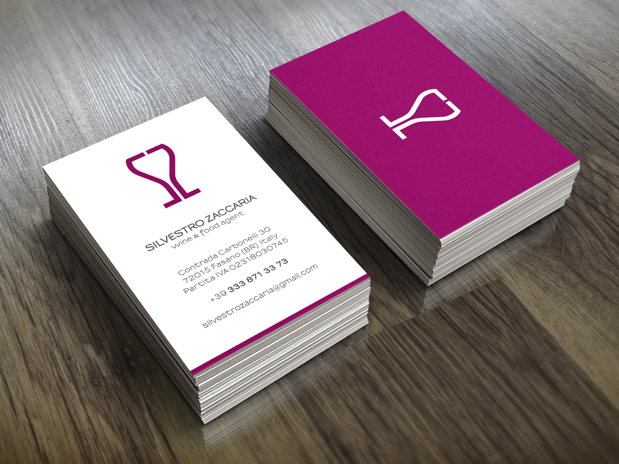 Mock-up of stacked Silvestro Zaccaria business cards in magenta and white, showing his S and Z monogram shaped like a wine glass, on a wooden table