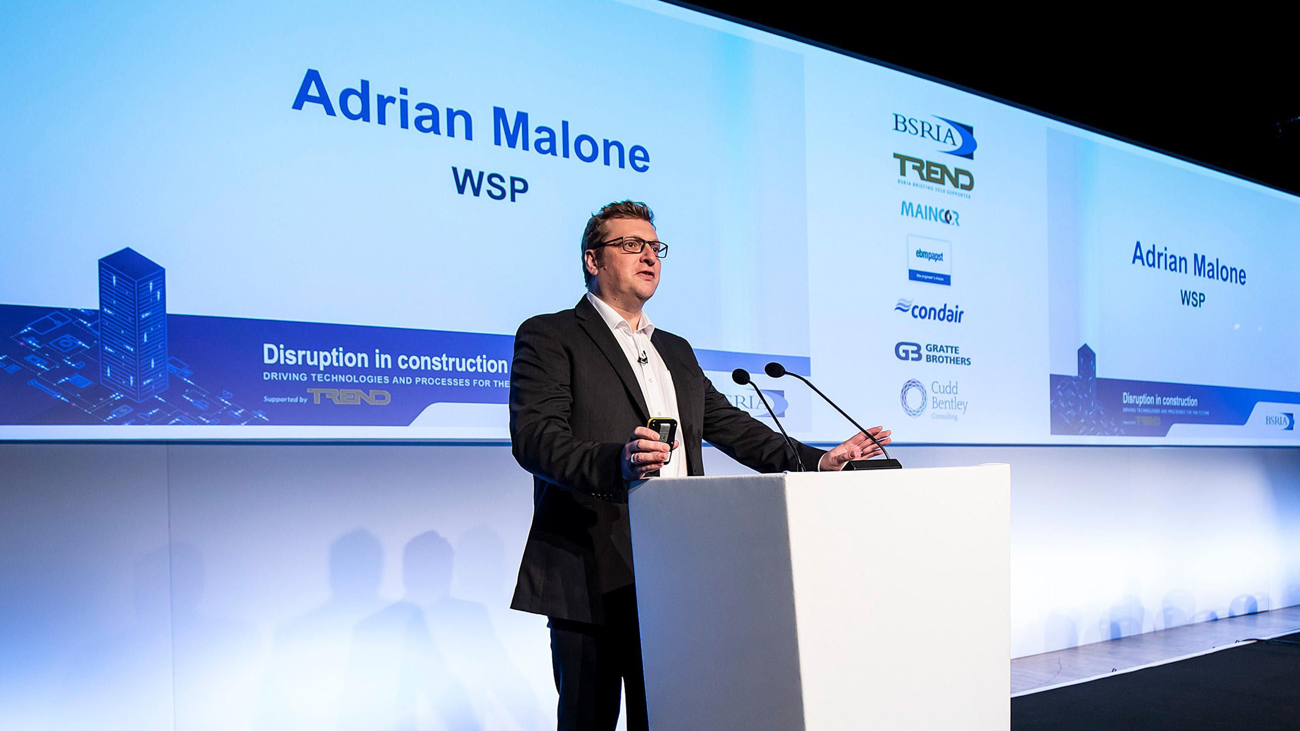 Speaker Adrian Malone of WSP presenting at the lectern in front of the 'Disruption in construction' stage graphics