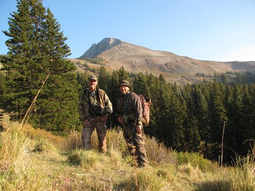 Two men in camouflage hiking gear stand in grass with pine trees and a rocky mountain in the background.