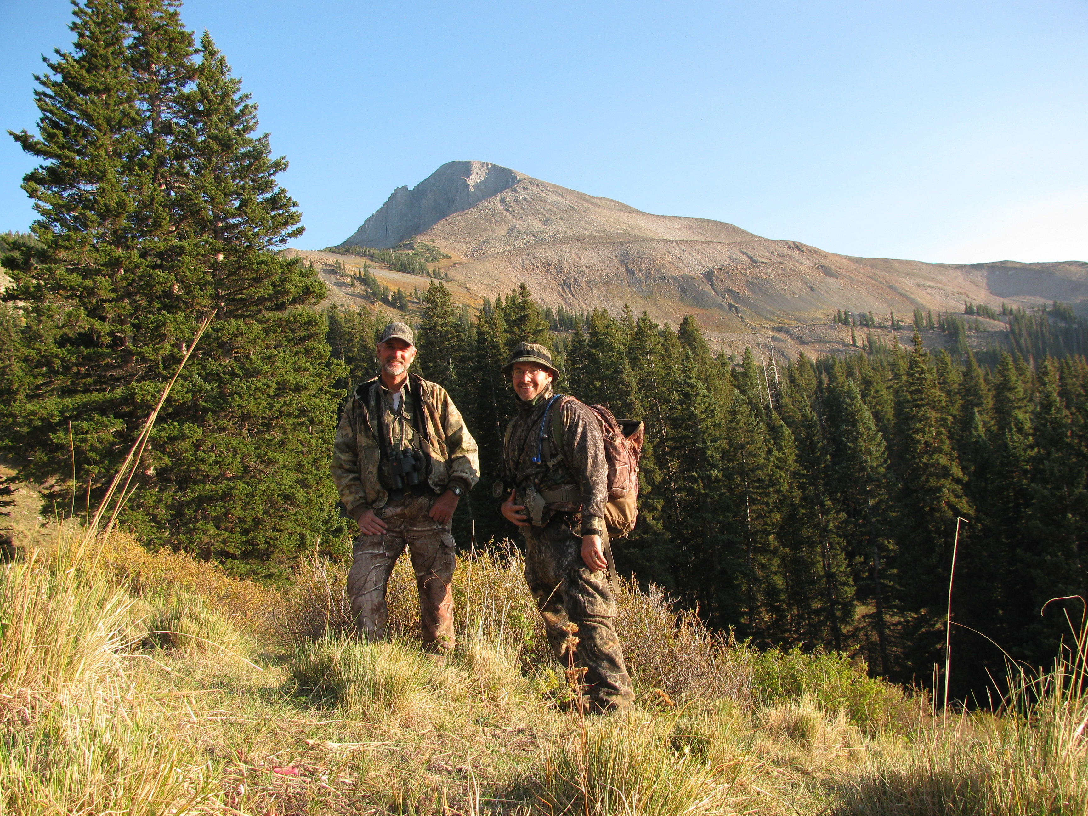 Two men in camouflage hiking gear stand in grass with pine trees and a rocky mountain in the background.