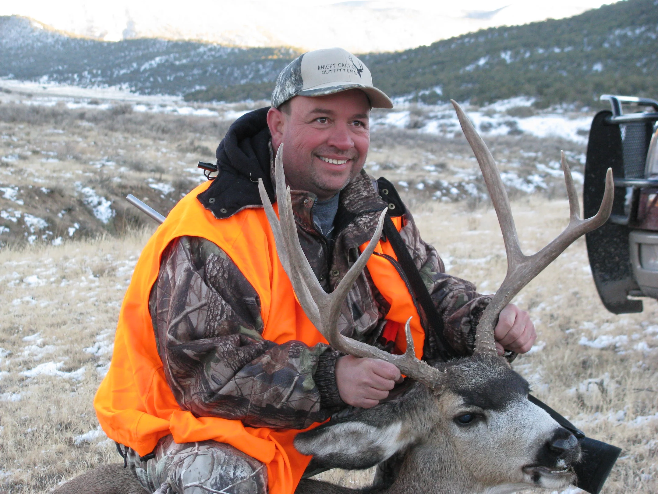 Smiling hunter in camo and orange vest holding a mule deer buck by its antlers in snowy terrain.