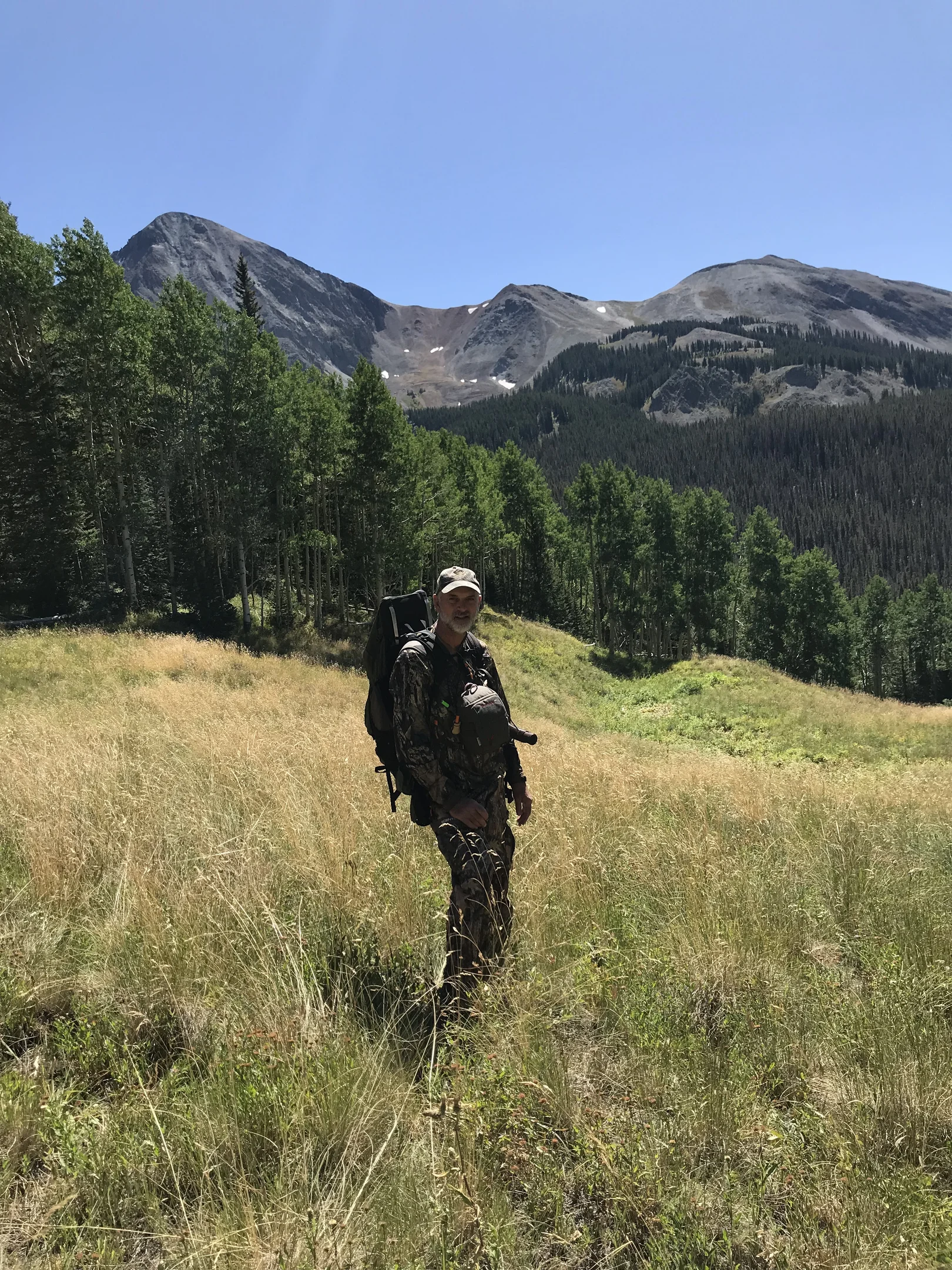 Hunter in camouflage with backpack standing in an alpine meadow with forest and mountains behind him.