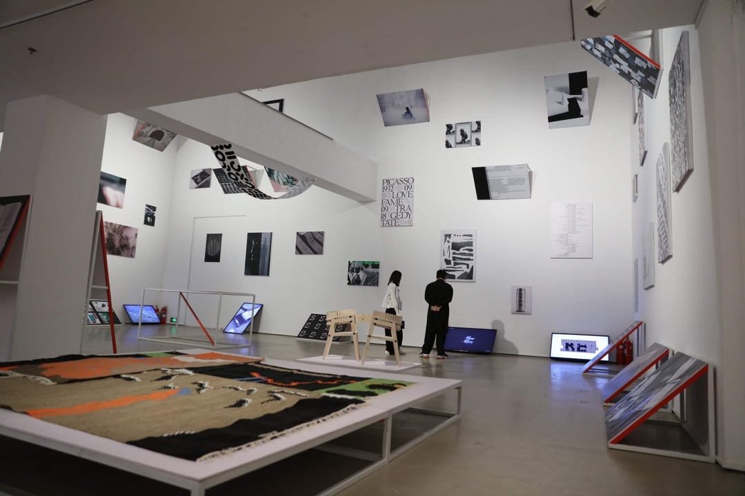 Two people viewing modern abstract art pieces displayed on white walls and platforms in a spacious gallery.