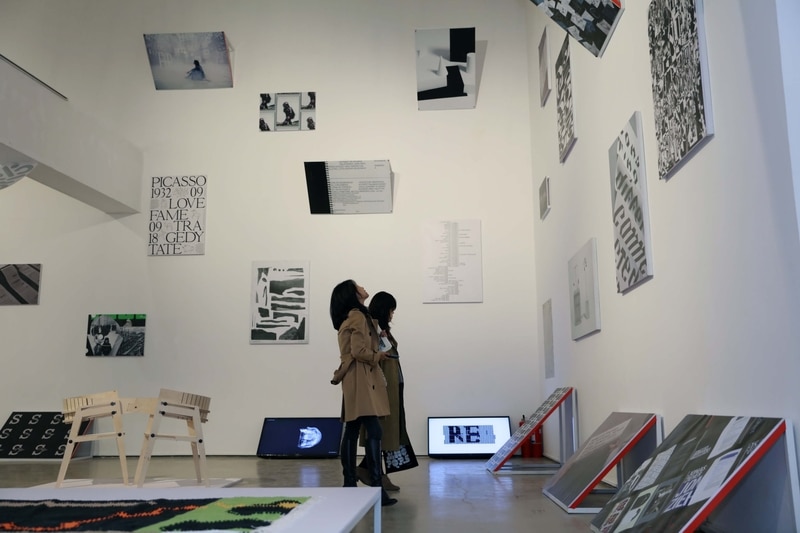 Two women in coats observing modern art pieces displayed on white walls and floor in a gallery.