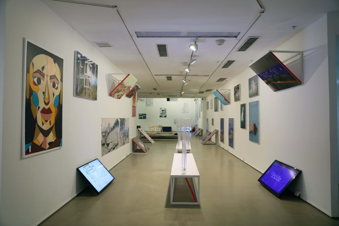 Interior of a contemporary art gallery with paintings and digital screens displayed on white walls and benches with clear sculptures in the center.