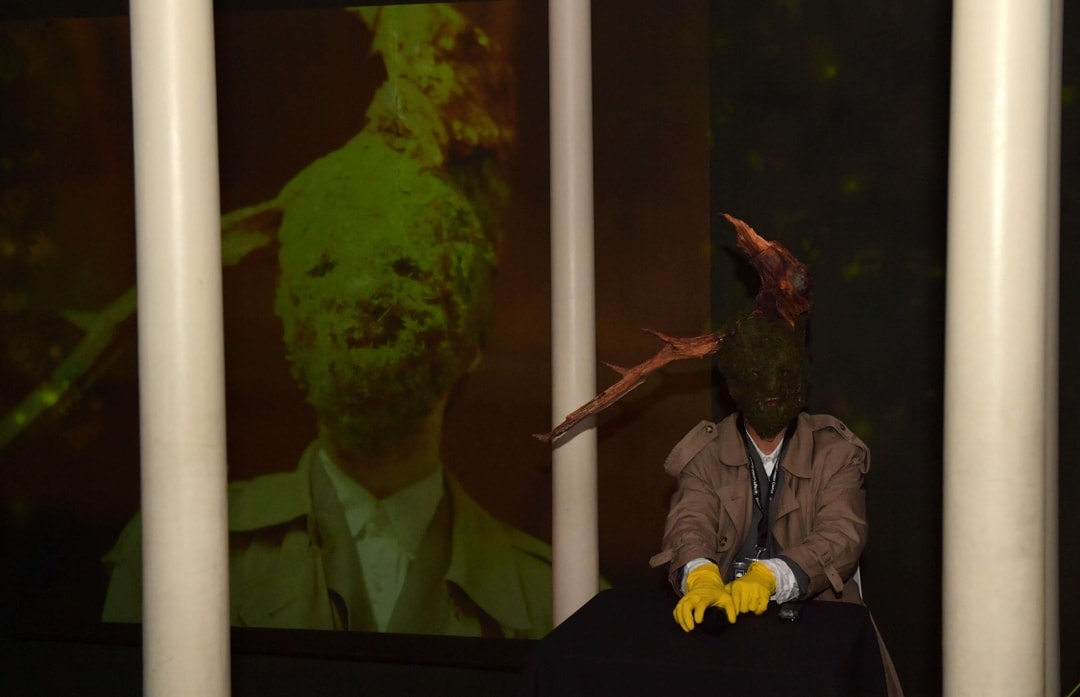 Person wearing a trench coat, yellow gloves, and a mask covered with moss and branches, sitting behind a black table with a dark background projection of the same masked figure.