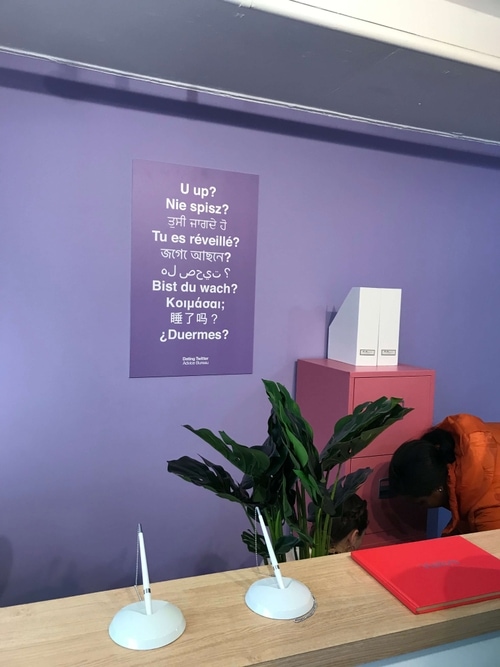 Office reception desk with two chained pens, a red visitor book, a large green plant, and two people near a pink filing cabinet against a purple wall with a multilingual poster asking 'Are you awake?'
