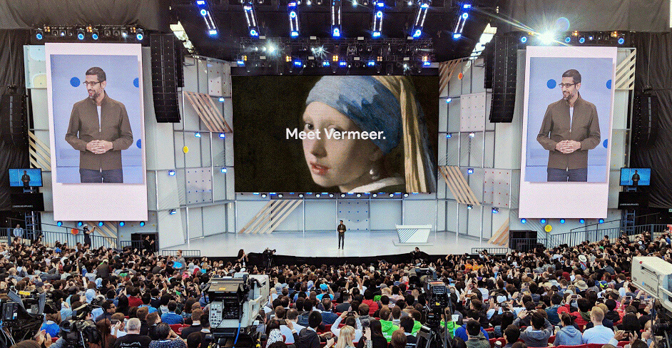 Presenter on large stage at a conference with two side screens showing him and a central screen displaying Girl with a Pearl Earring painting and text 'Meet Vermeer.'