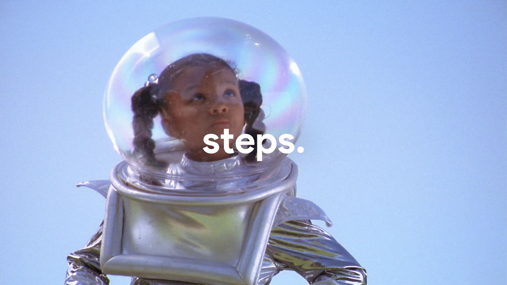 Child in a shiny silver astronaut costume with a clear bubble helmet looking up against a blue sky.