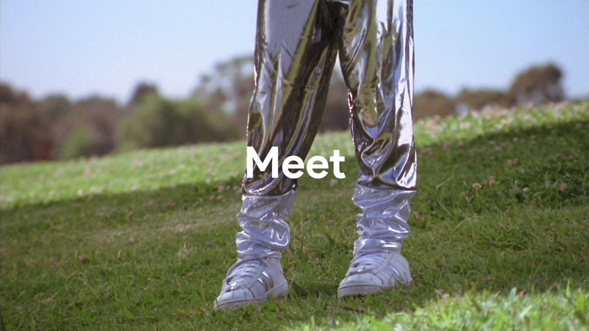 Person standing on green grass wearing shiny silver pants and white sneakers with the word 'Meet' overlaid in white text.