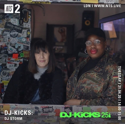 Two women sitting behind a counter covered with stickers, one wearing glasses and a camouflage jacket, the other with straight hair and a black fur vest.