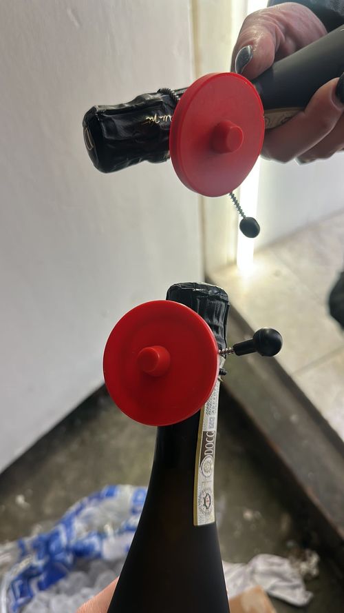 Two black bottles with foil tops, each fitted with a red plastic safety cork stopper, one held by a hand with painted nails.