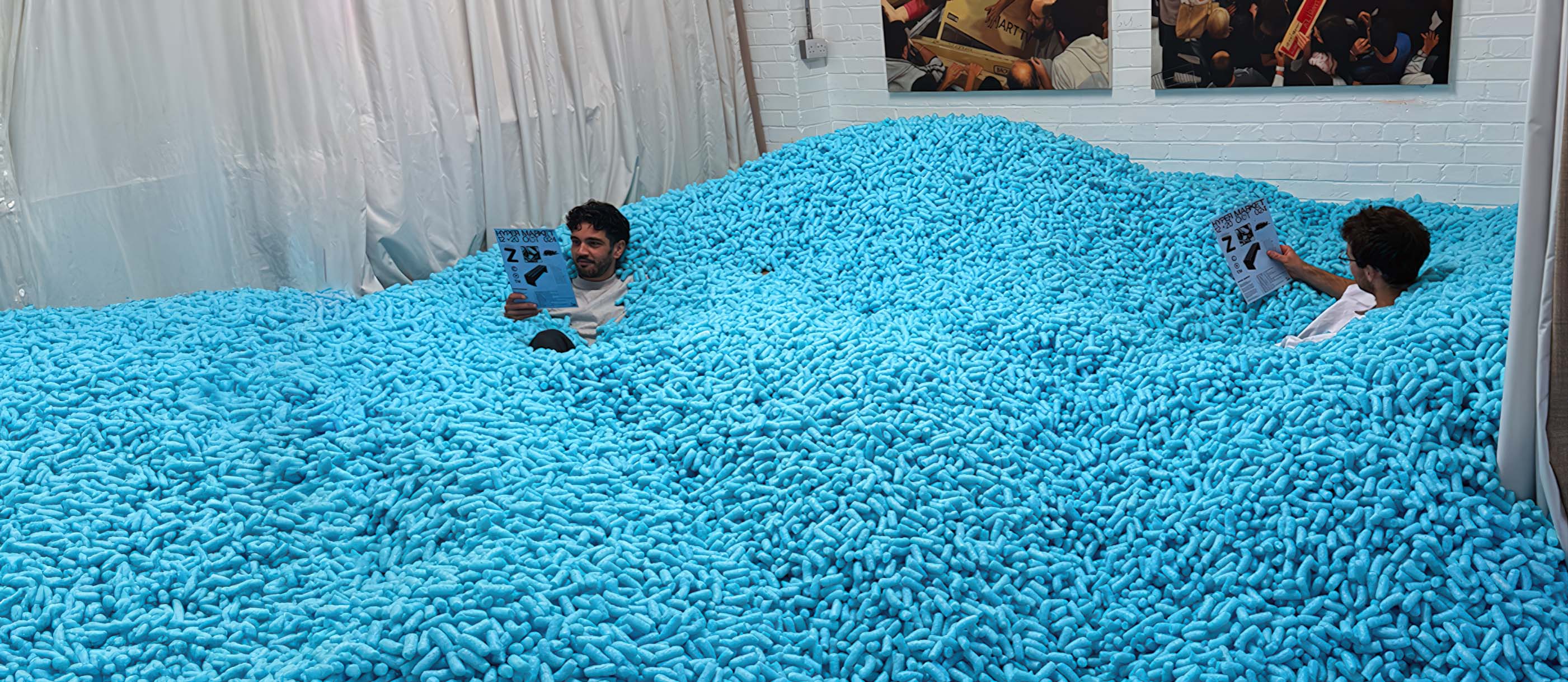 Two men partially submerged in a large pit filled with thousands of blue packing peanuts, each reading a sheet of paper.