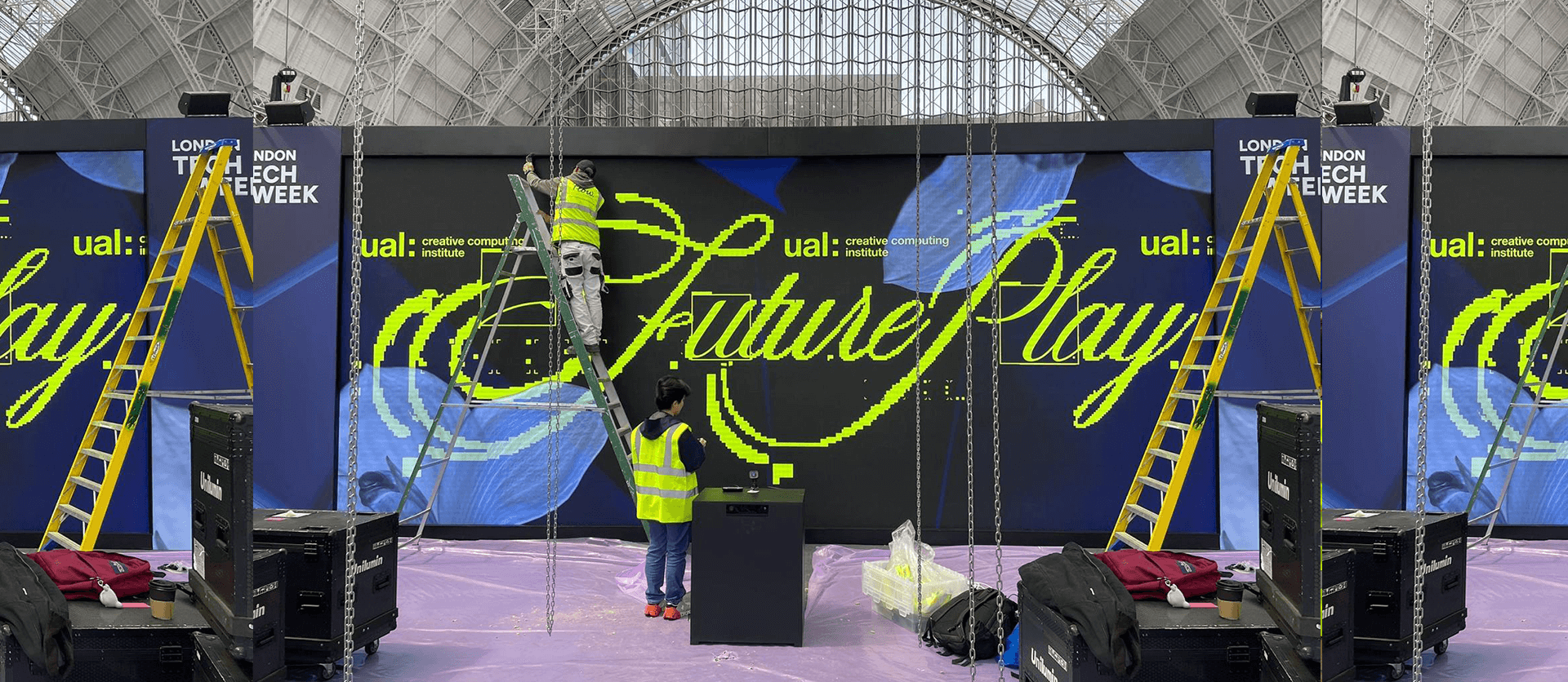 Two workers in high-visibility vests setting up a large LED display screen with 'Future Play' text at London Tech Week event inside a large glass-roofed venue.