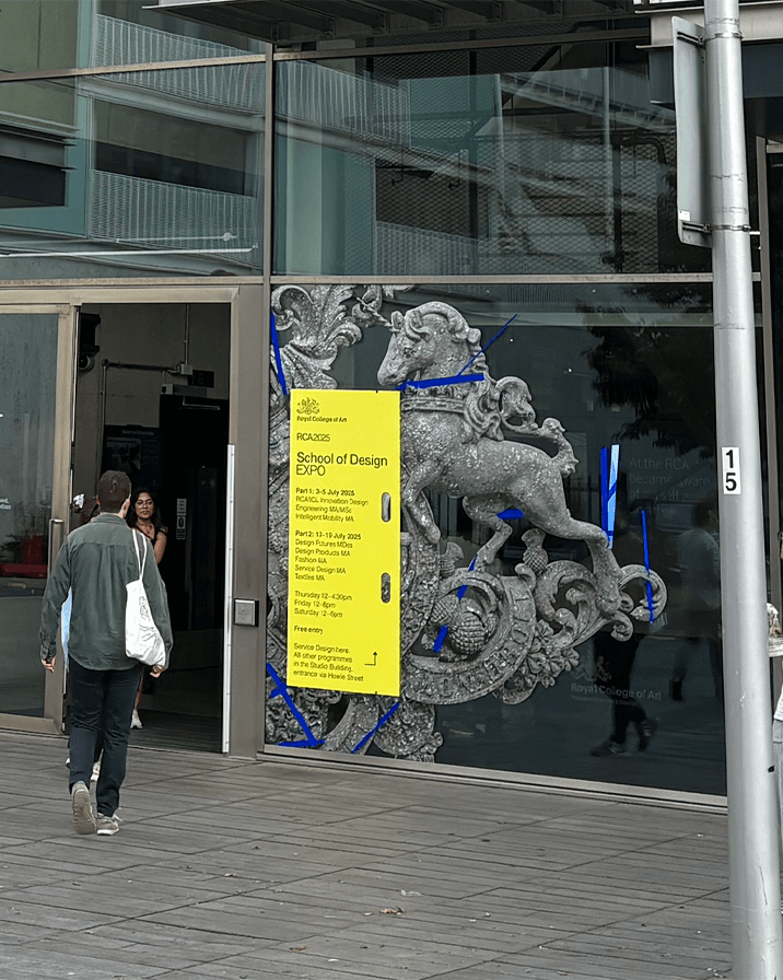 Exterior glass wall of a building featuring a large decorative stone lion relief and a bright yellow poster for the Royal College of Art's School of Design Expo with schedule details, while a person carrying a white tote bag walks inside.