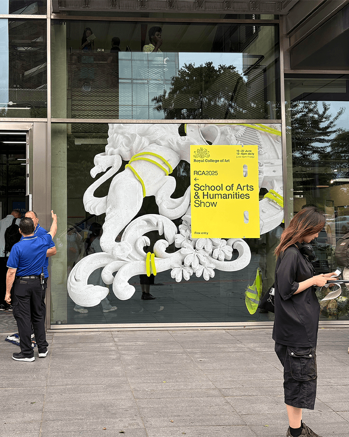 Entrance window display of Royal College of Art featuring a large white decorative sculpture with yellow cords and a yellow sign for School of Arts & Humanities Show.