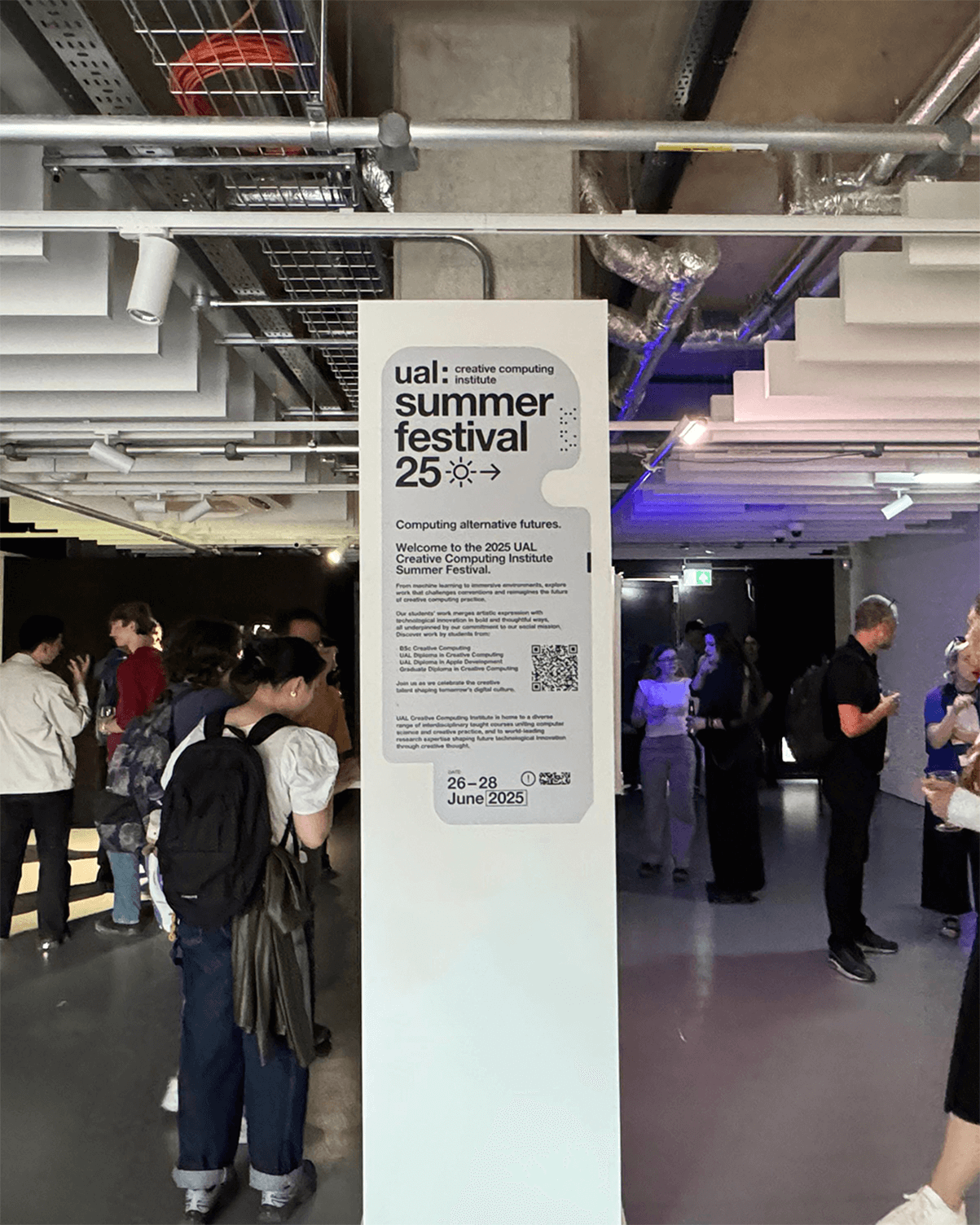 Indoor event space with people socializing around a tall white sign for the UAL Creative Computing Institute Summer Festival, dated 26-28 June 2025.