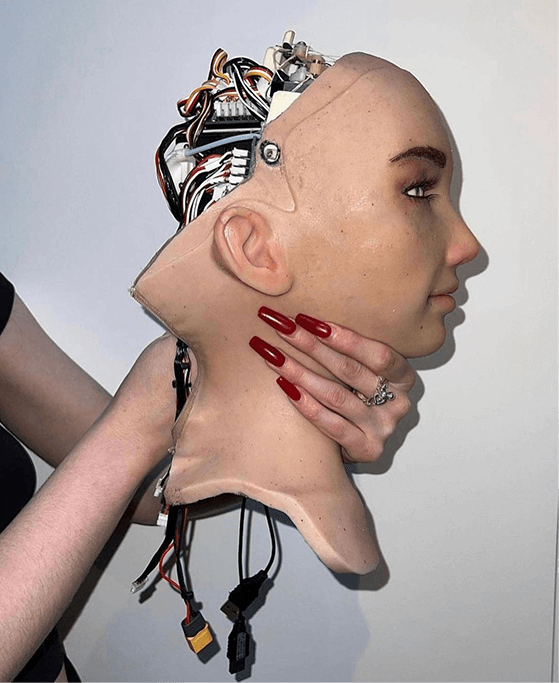 Person with long red nails holding a realistic female robot head with exposed wiring and connectors at the back.