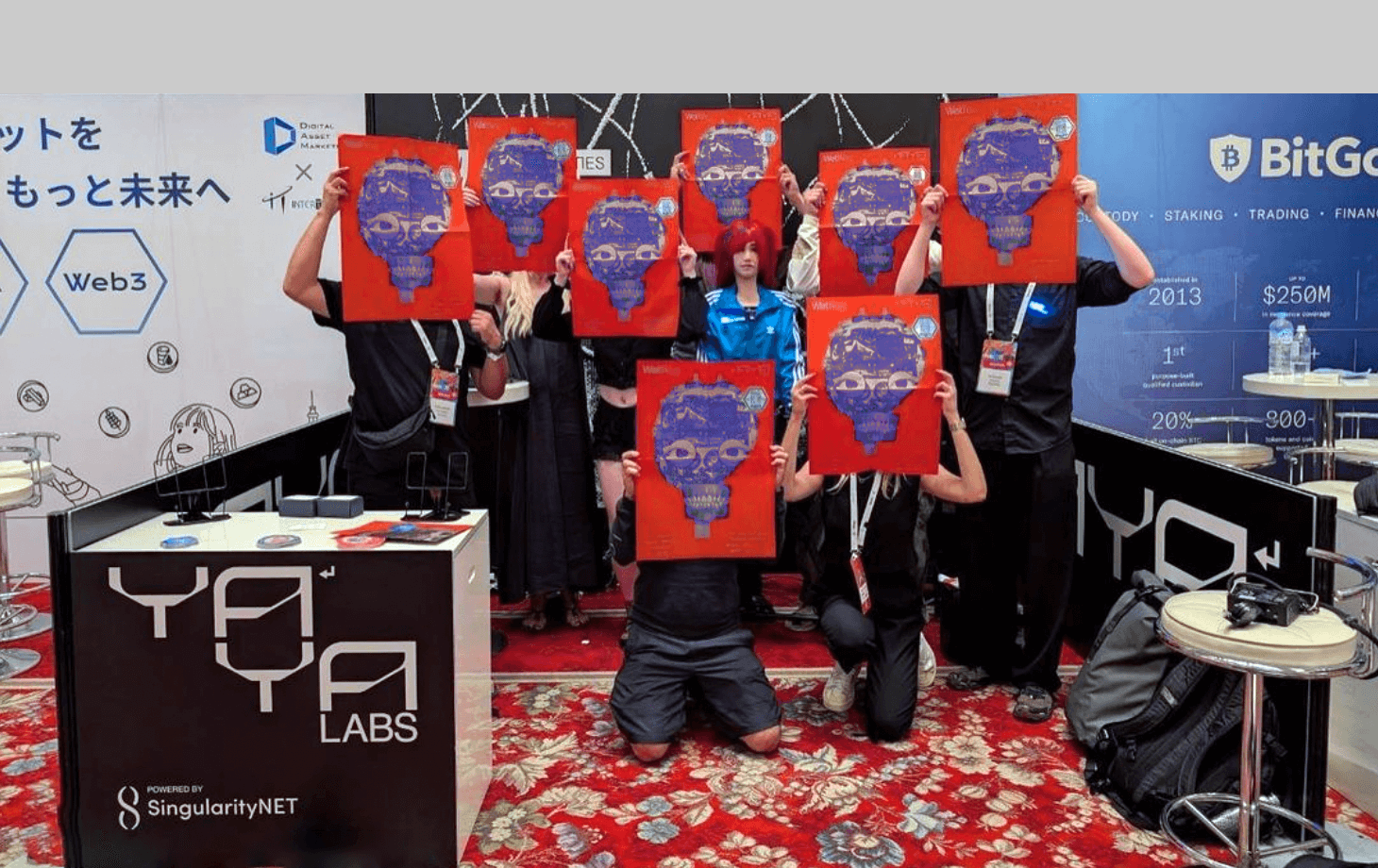 Group of eight people at a conference booth, seven holding up red posters with blue skull designs covering their faces, standing behind a YA Labs branded counter.