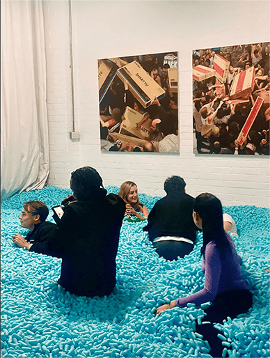 Five people sitting and lying in a room filled with numerous small blue foam cylinders, with two photos of crowded scenes on the white brick wall behind them.