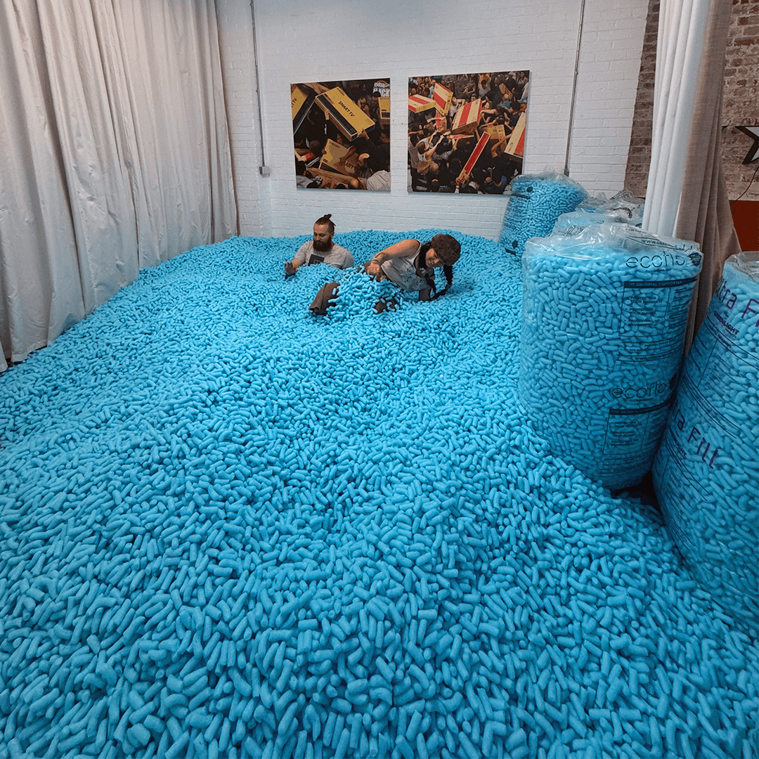 Two people sitting and lying in a large indoor pit filled with blue foam packing peanuts, with large bags of the peanuts stacked on the side.
