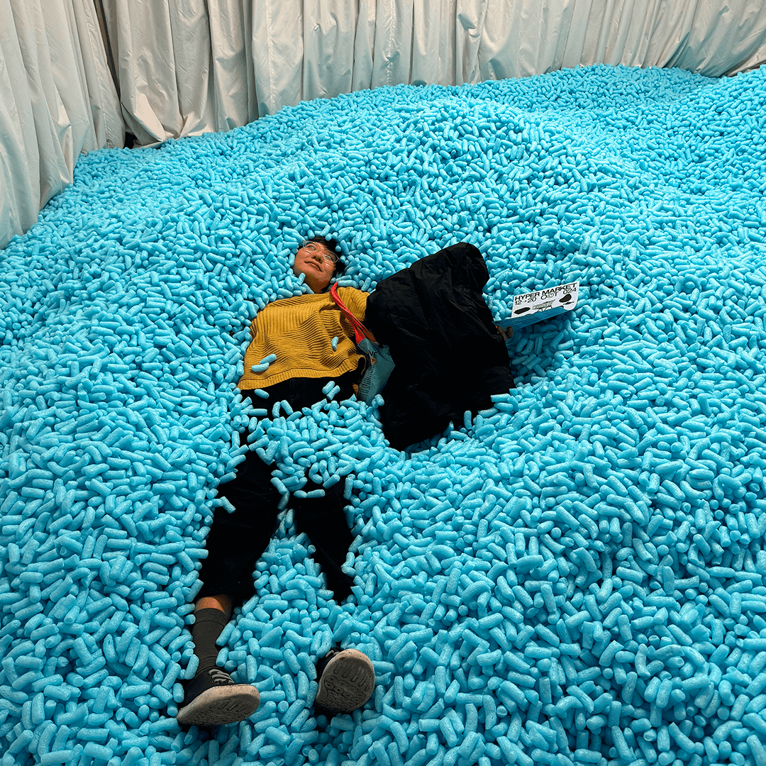 Person lying on their back in a large pool filled with blue foam packing peanuts, holding a sign and partially covered by a black jacket.