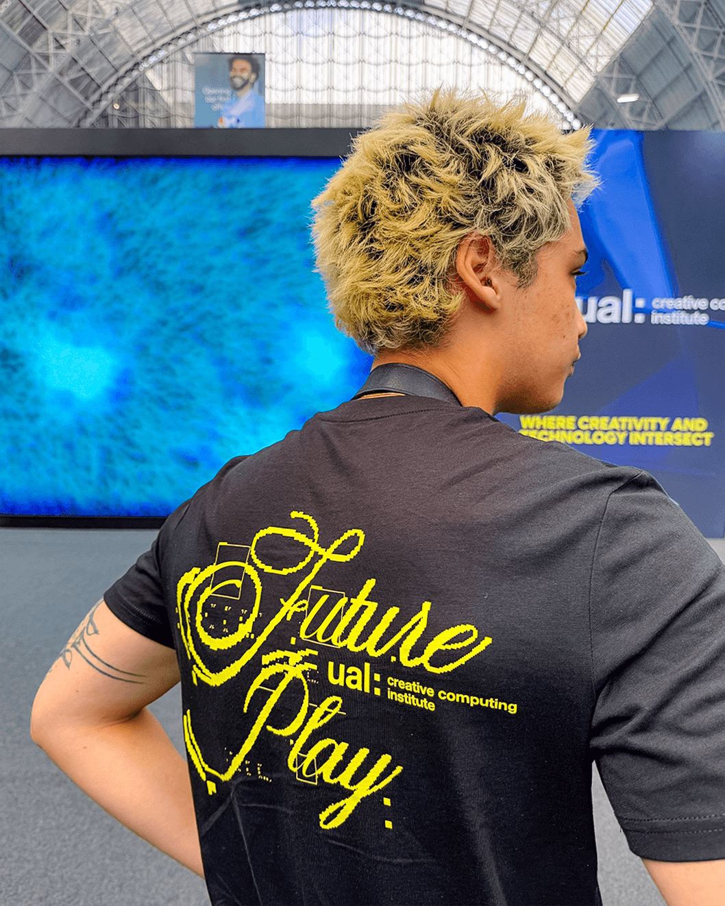 Person with blond curly hair wearing a black T-shirt with yellow text reading 'Future Play' and 'ual: creative computing institute' on the back inside a large indoor space with blue digital displays.