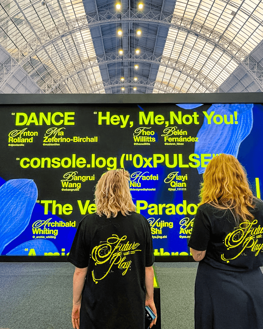 Two people with 'Future Play' printed on black shirts stand in front of a large digital display with bright yellow-green text and blue shapes under a glass arched ceiling.